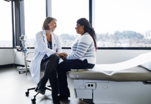 Smilende lege snakker med pasient som sitter på en sykehusseng.